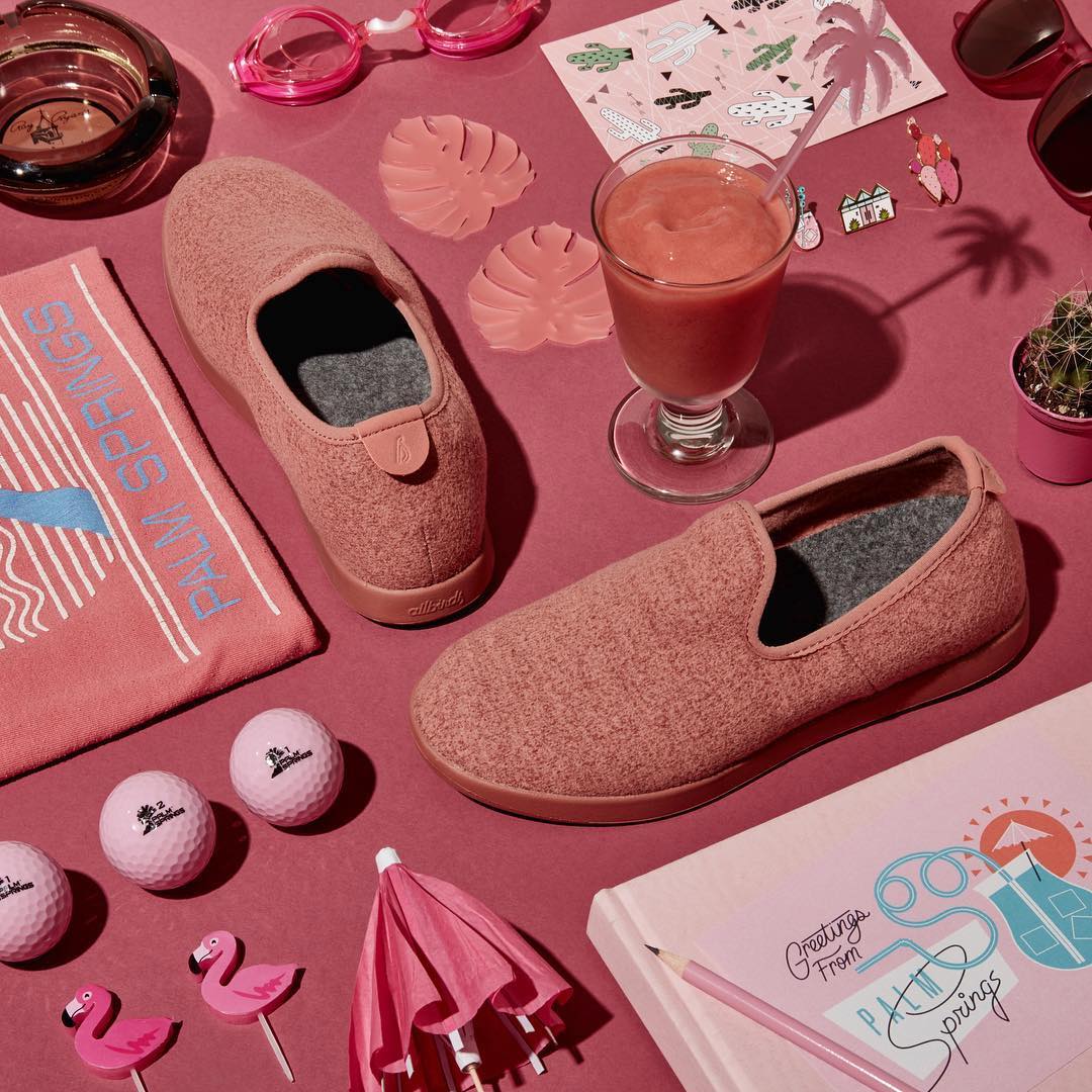 allbirds wool loungers in pink in a flat lay with other pink items including a cocktail umbrella, books, and a beverage