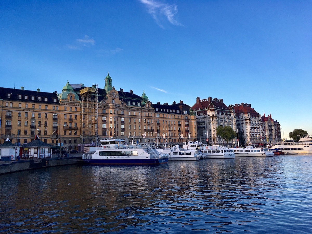 A view of buildings in Stockholm. They are about 7 to 8 stories tall, with ornate architecture, and by a body of water. There are small boats docked in the water.