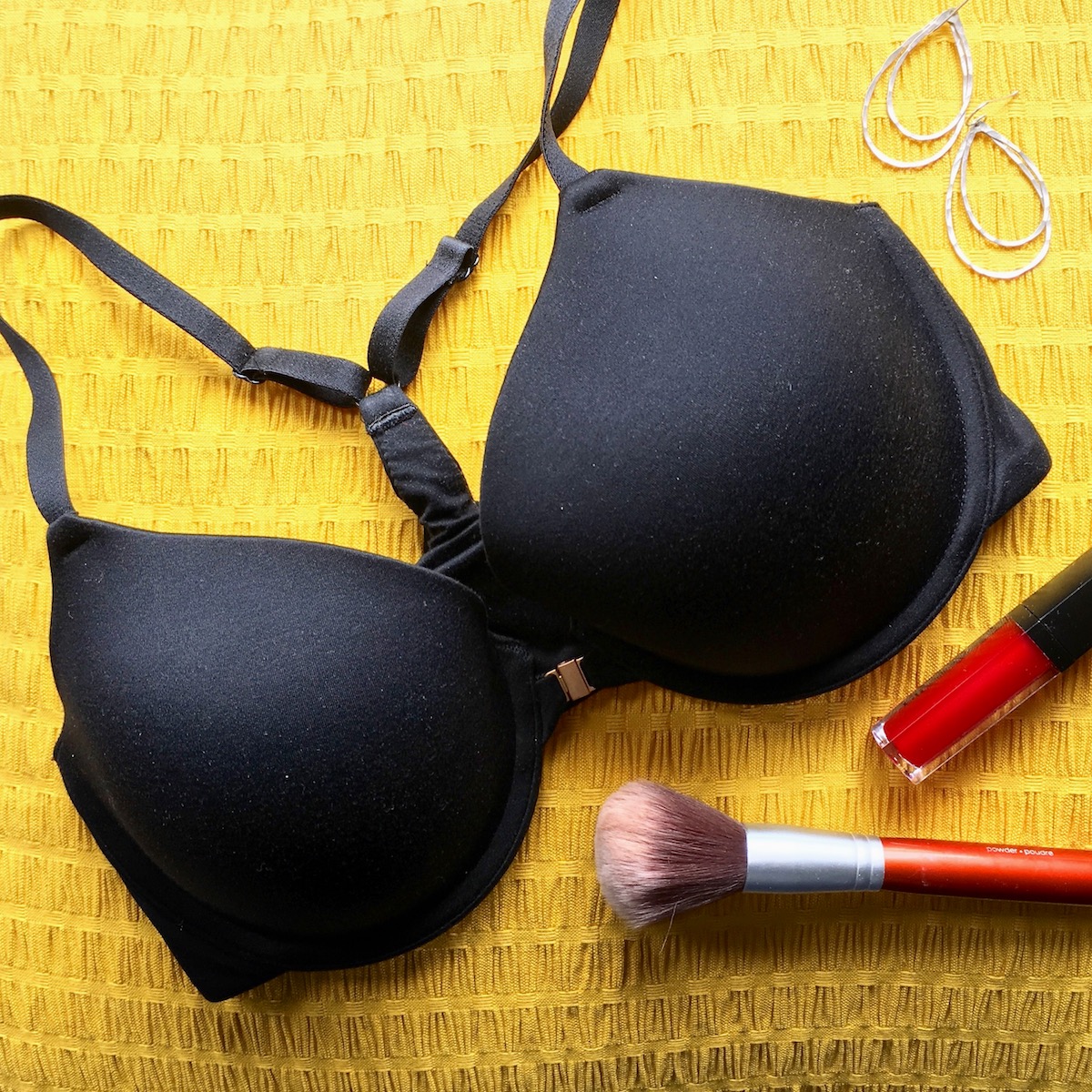 Flat lay photo of a black racerback bra with silver earrings, a makeupbrush, and red lip gloss.