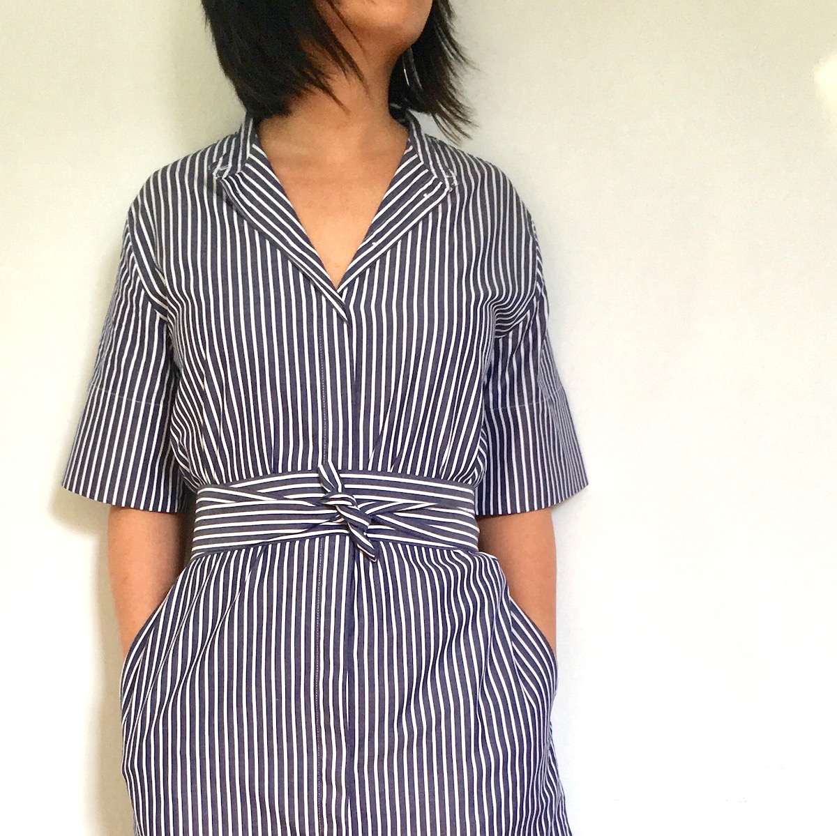 A woman with black hair wearing the striped Everlane cotton collarless belted shirt dress. Her hands are in the pockets.