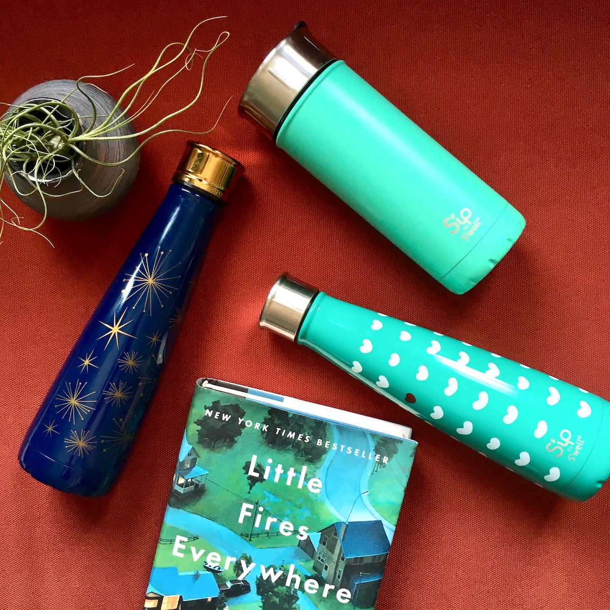 Three S'ip by S'well water bottles laying against a red background, next to a book and a plant. The book is "Little Fires Everywhere" by Celeste Ng.
