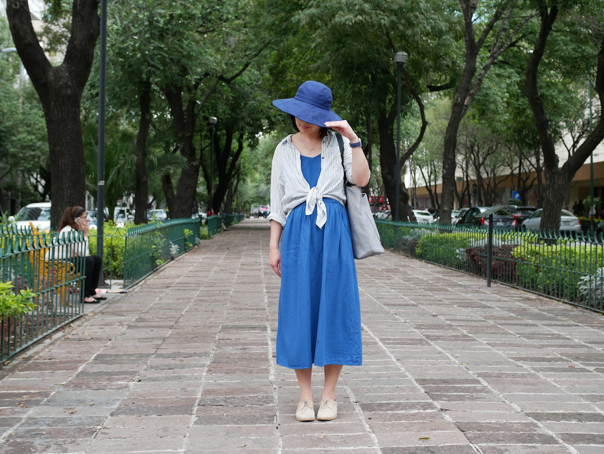 How to pack light for vacation: A woman stands on a brick walkway. She's wearing a mid dress, a long sleeve shirt tied at the front, and a hat.