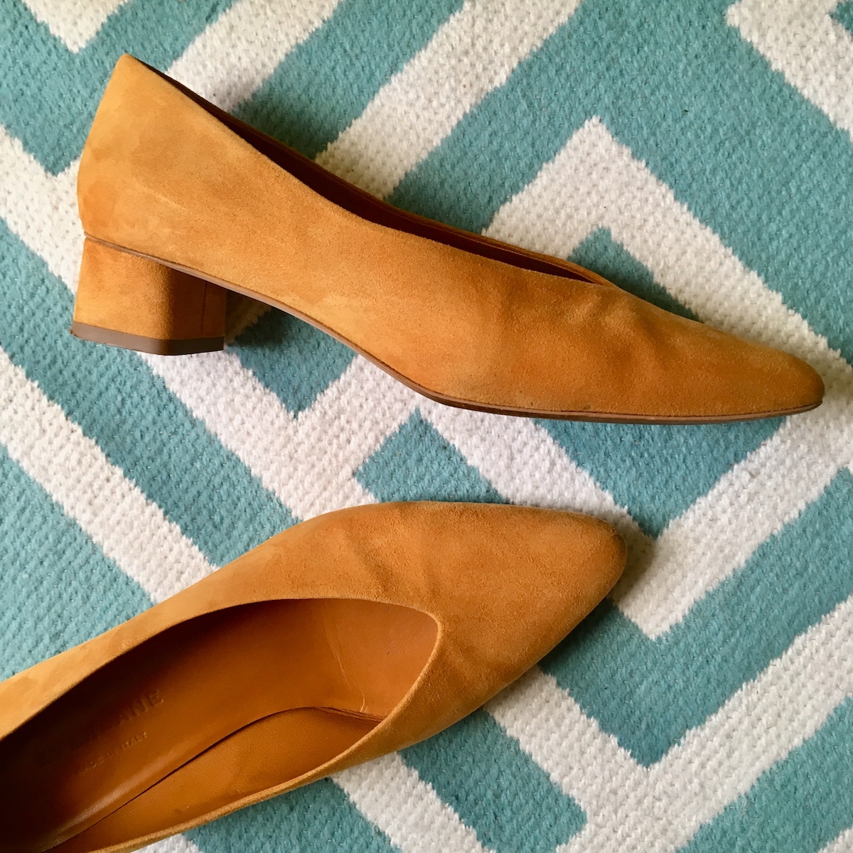 A pair of mustard colored suede Everlane V heels lay on a blue and white rug.