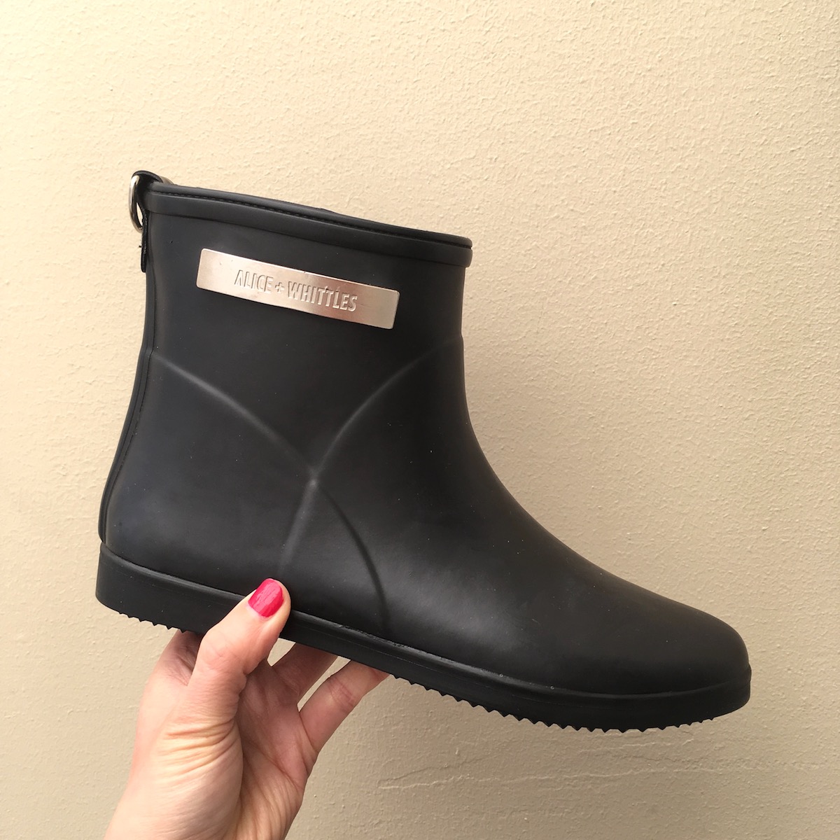 Alice + Whittles ethical rain boot review: A hand with red nail polish holds up a black rain boot. There is a metal plate at the top of the boot, which reads, "Alice + Whittles"