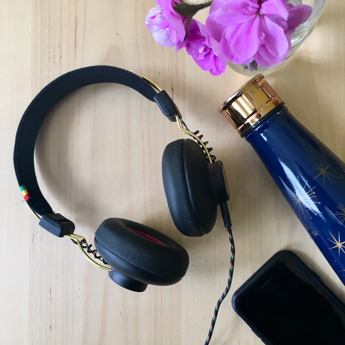House of Marley Positive Vibrations 2 headphone review: A pair of on-ear headphones on a wooden surface net to a stainless steel water bottle, a phone, and a purple flower in a jar.