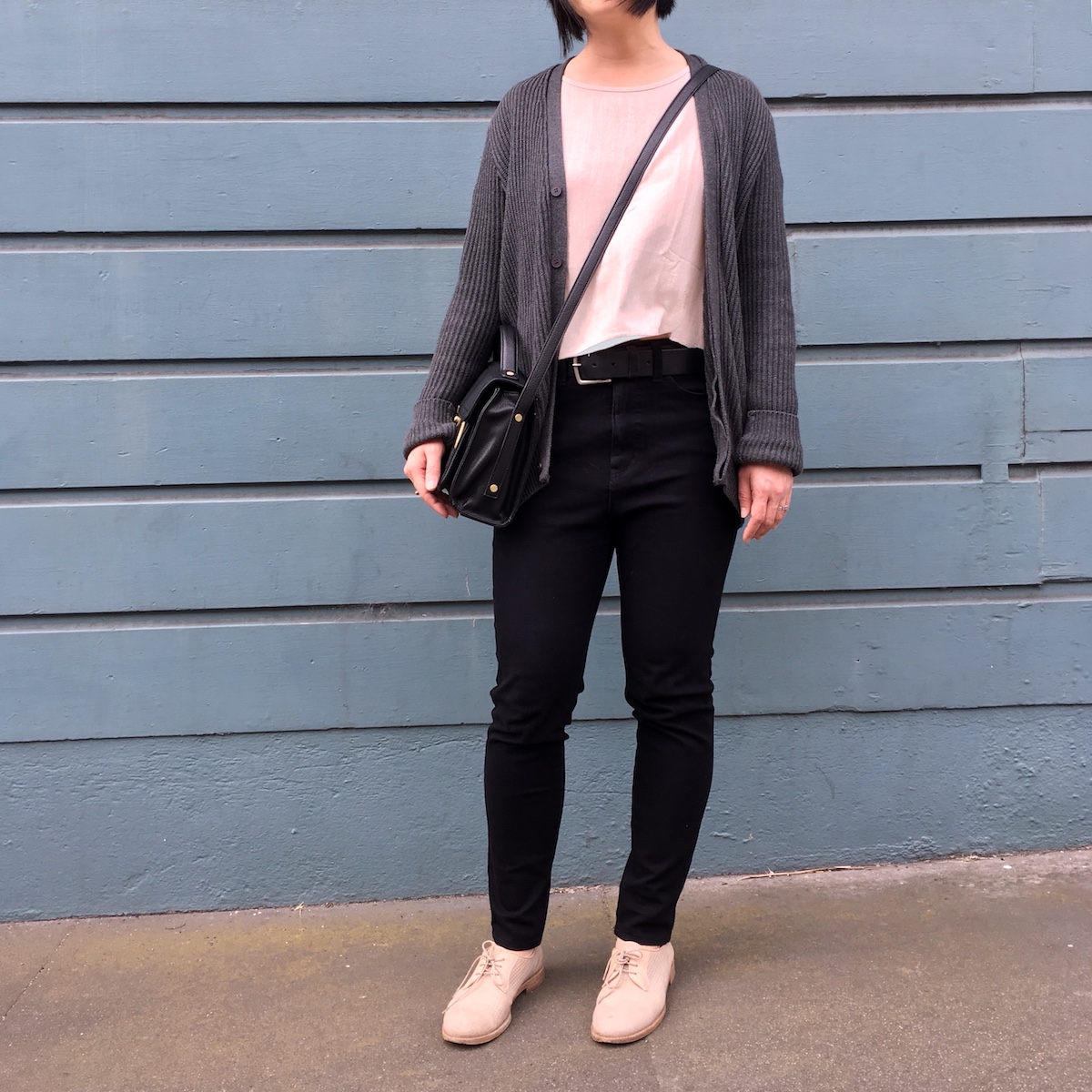 A woman, her head not in the frame, wearing an oatmeal-colored cropped shirt, black jeans, and a gray cardigan. She is also wearing a black purse cross body and is standing in front of blue siding.