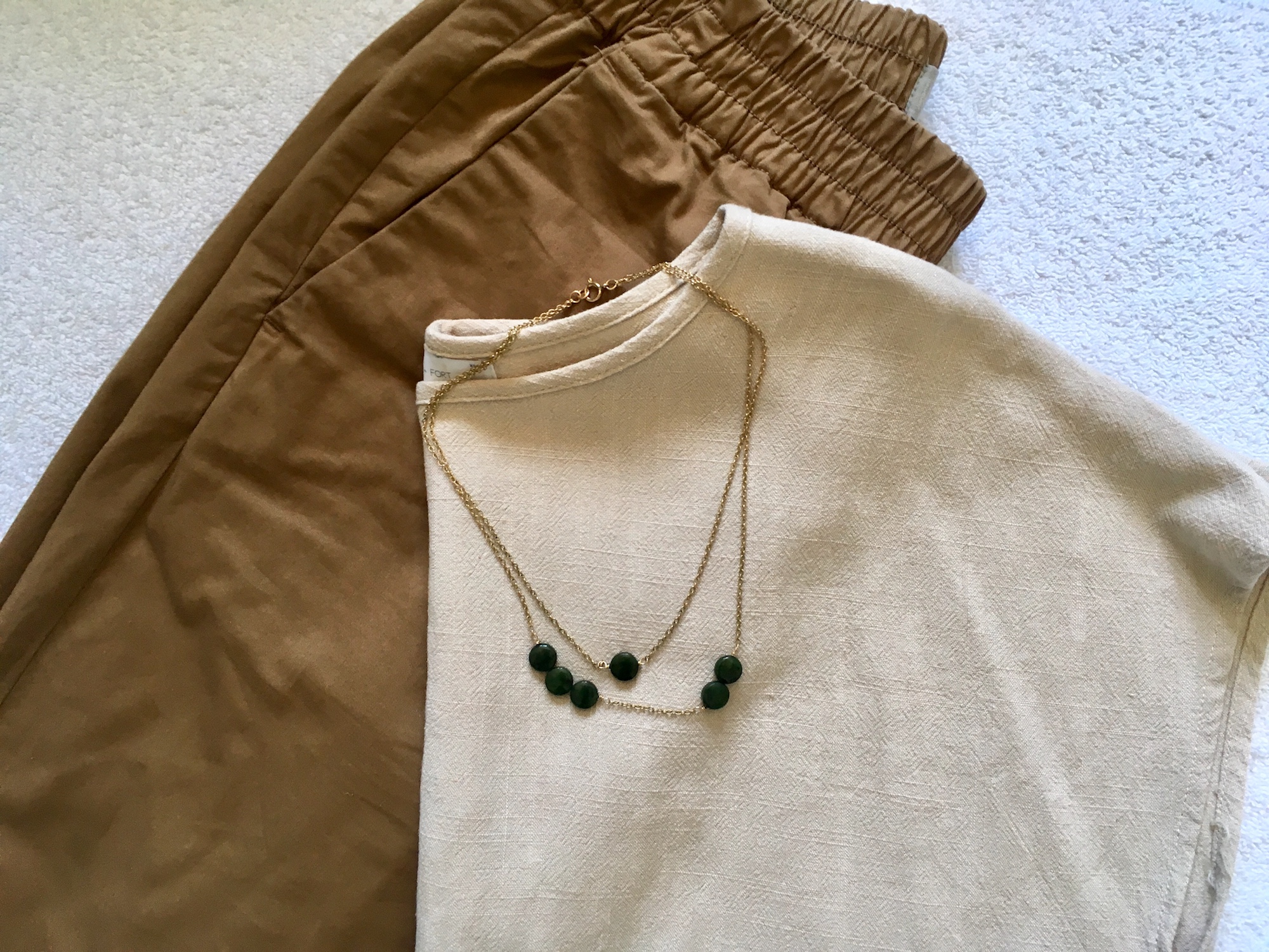 A ochre pants and a beige top lay on a white background. A gold necklace with green beads is on top of the shirt.
