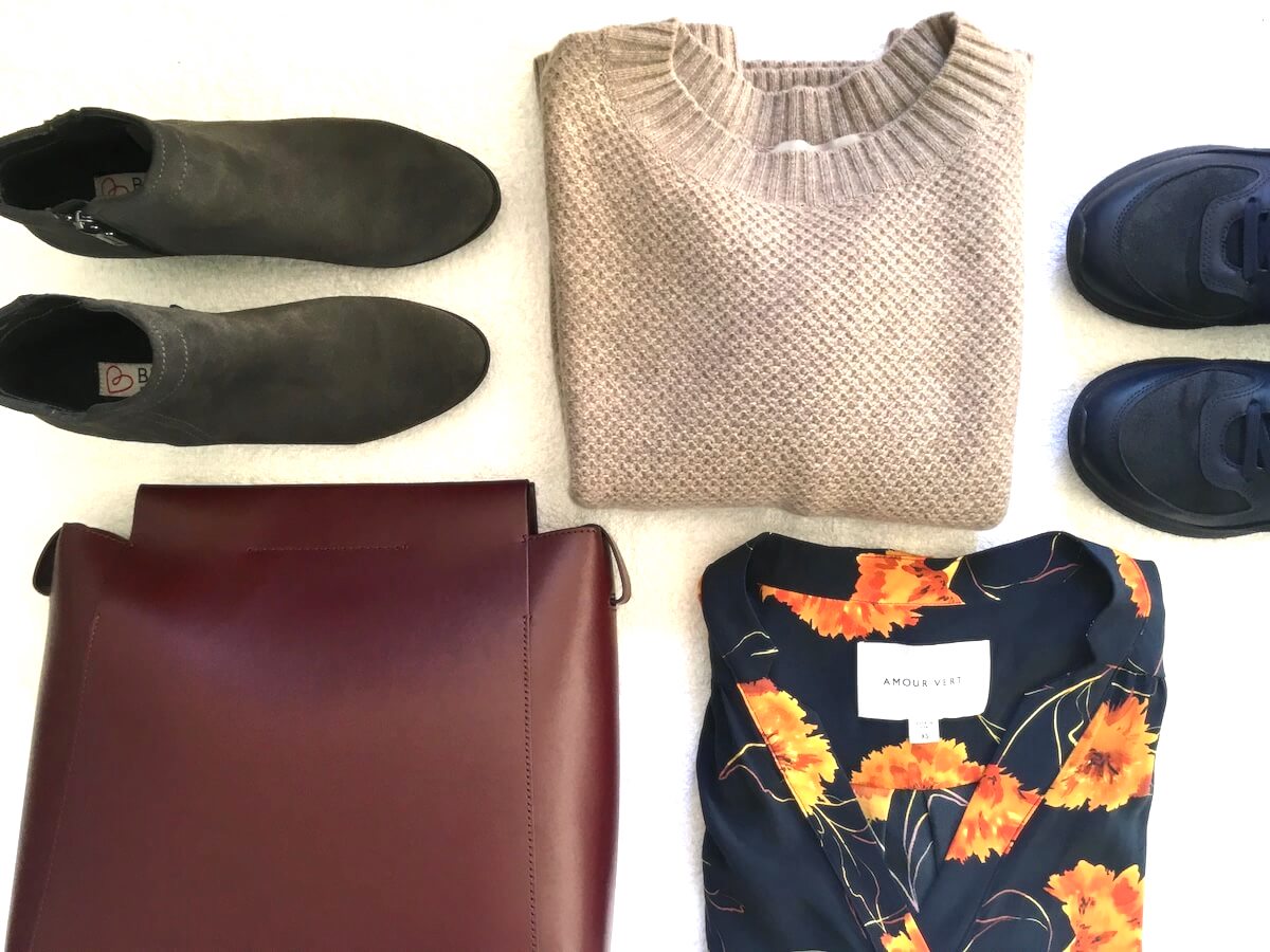 My Year in Shopping Less: a flat lay of clothing on a white surface, including gray boots, a brown sweater, navy sneakers, a floral print shirt, and a maroon leather bag.