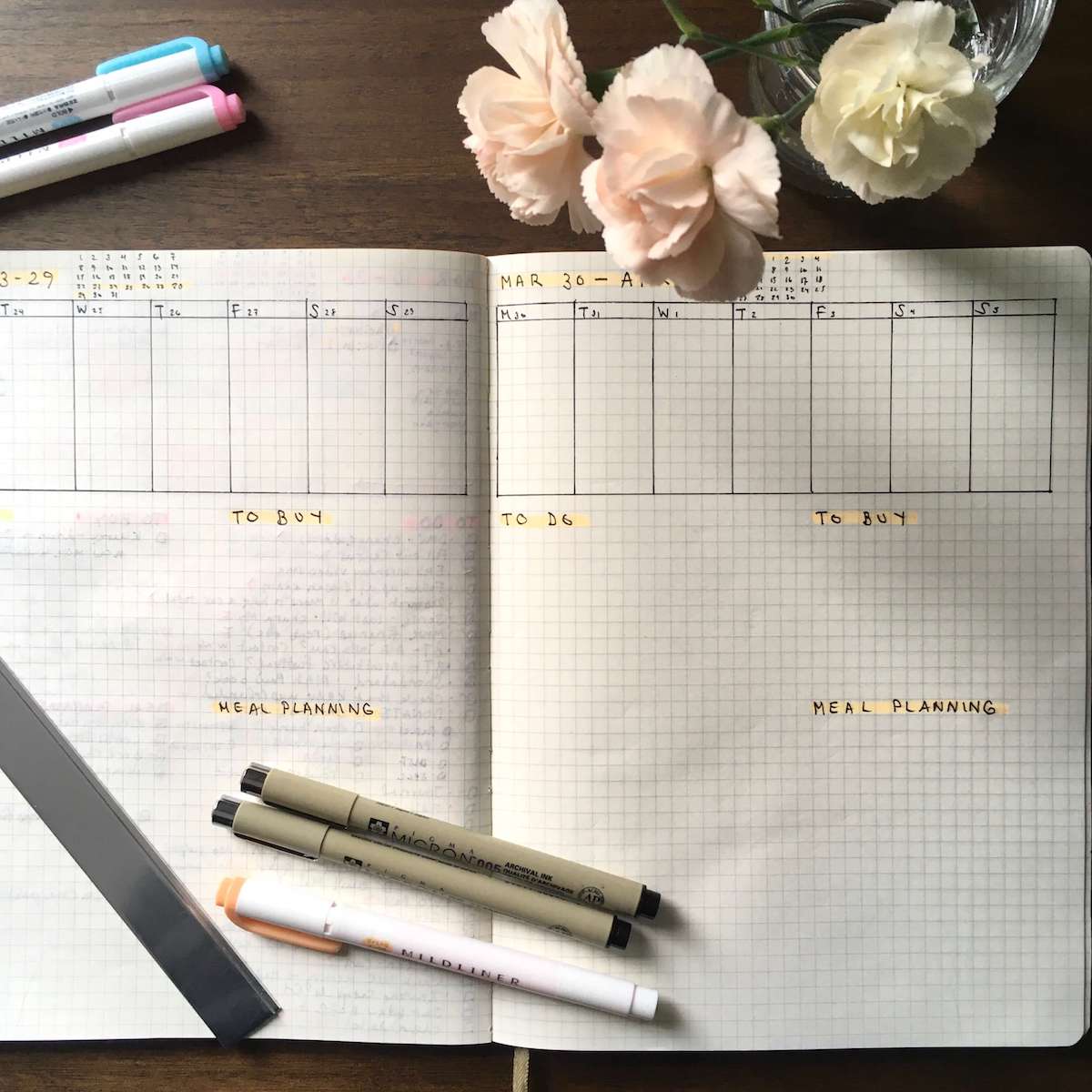 A bullet journal with a weekly view open on a desk. There is a small jar of flowers and pens.