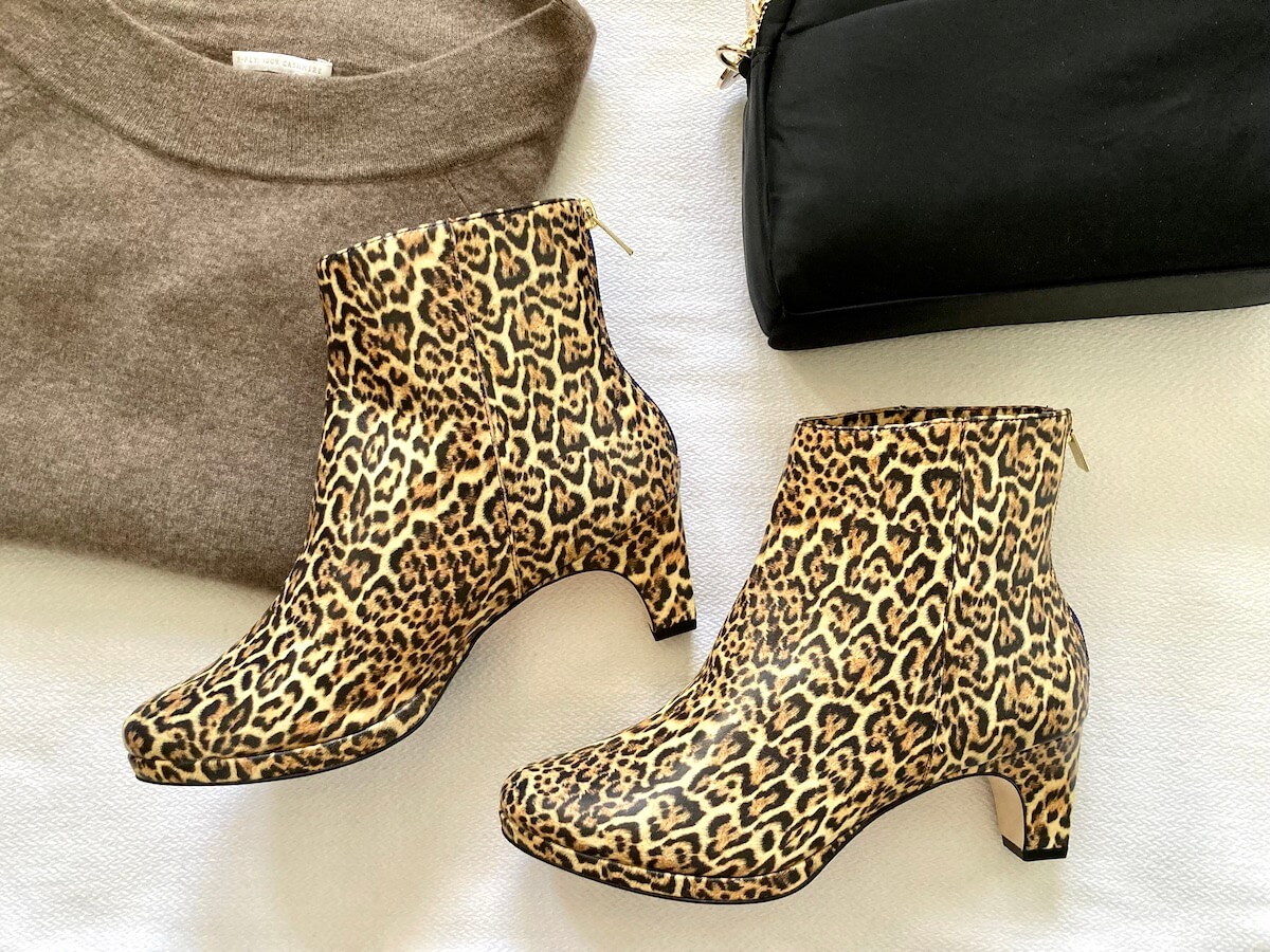 A pair of leopard print booties on a white surface, next to a folded sweater and a black bag.