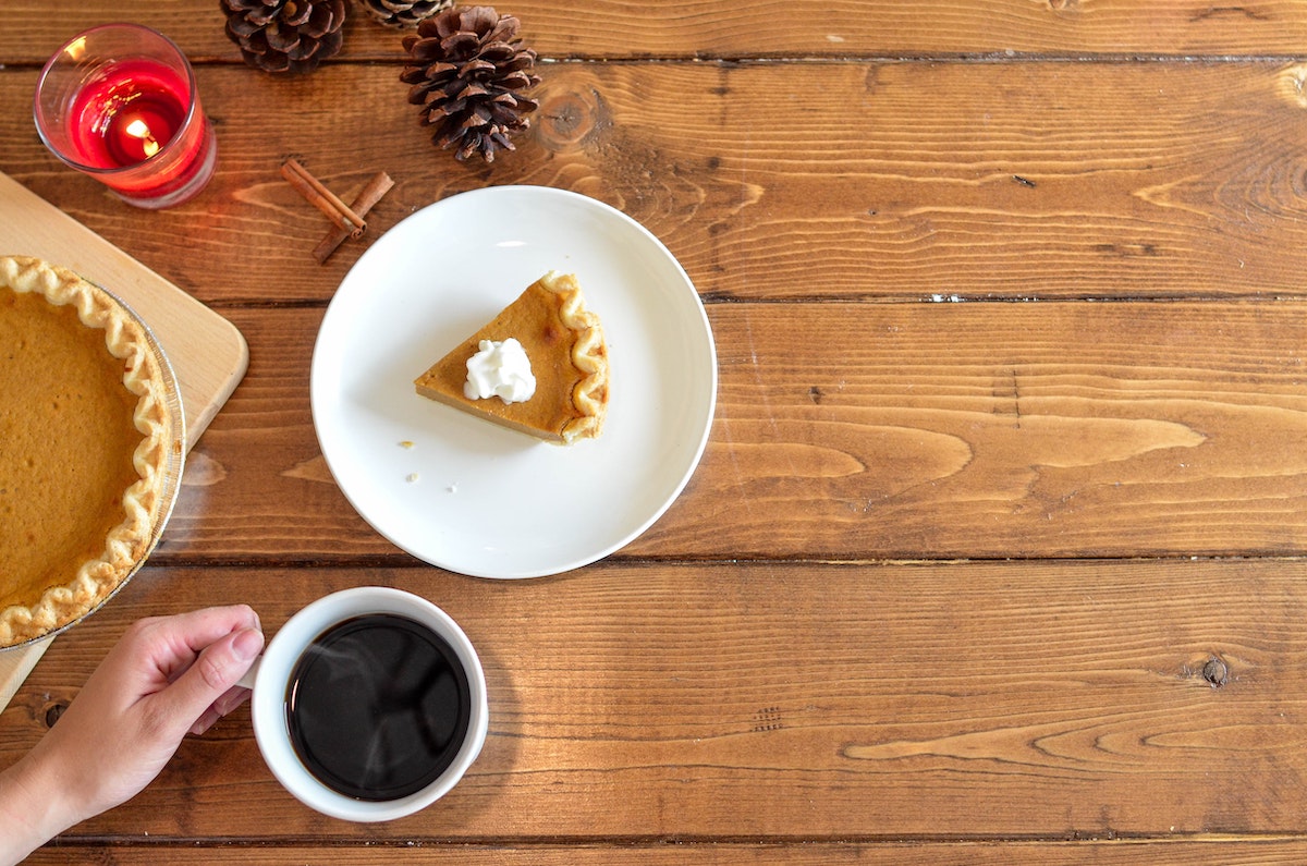 View from above a wooden table of a slice of pumpkin pie on a white plate and a hand holding a cup of black coffee.