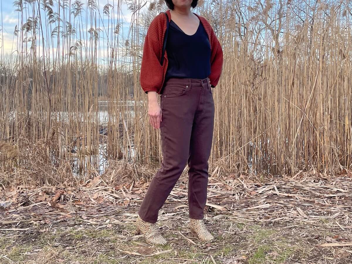 A person with black hair standing outside in front of tall straw plants, wearing the Everlane Cheeky Jean in brown with a red-orange cardigan.