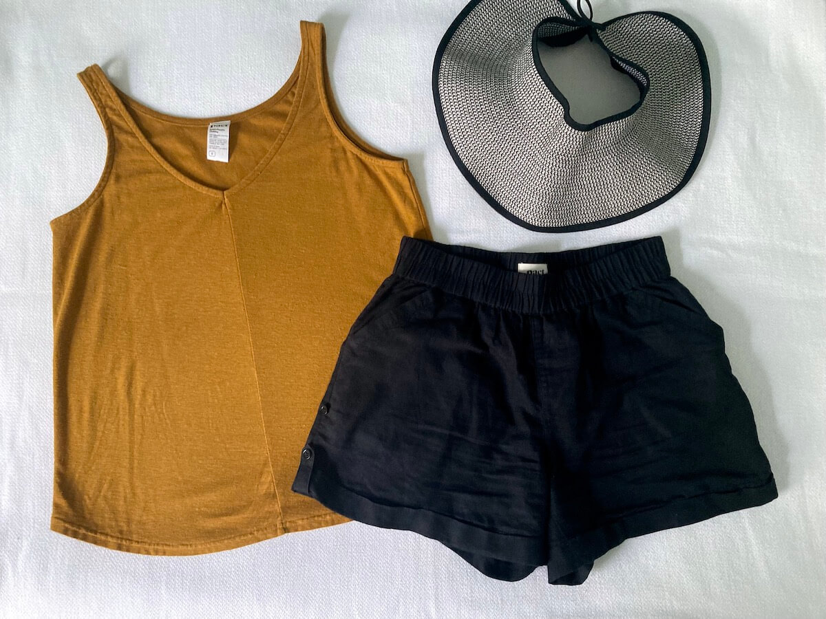 A flat lay of items on a white surface: a golden brown/yellow tank top, black shorts, and a black and white roll-up hat.