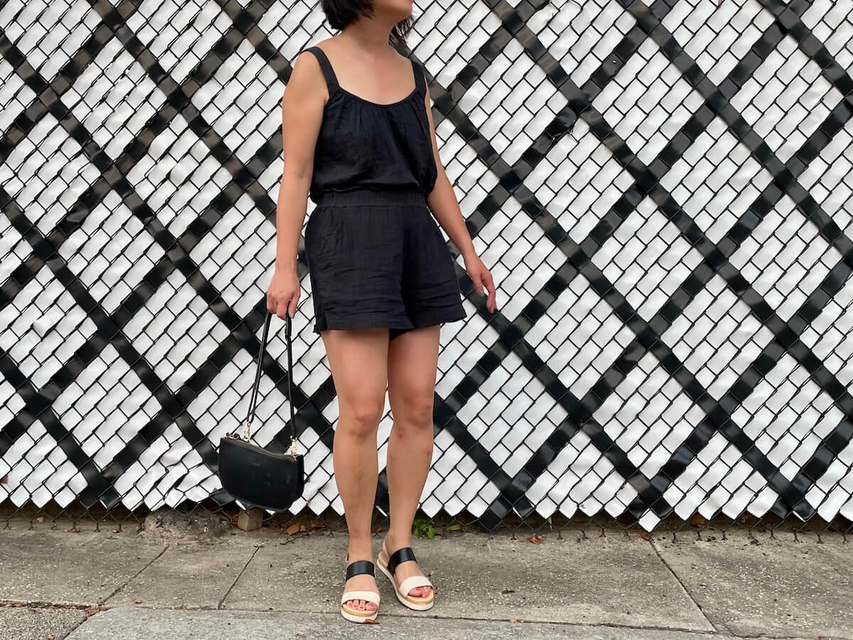 A person (me) with black hair wears a faux romper with a crew neck, posing for a photo in front of a white and black fence.