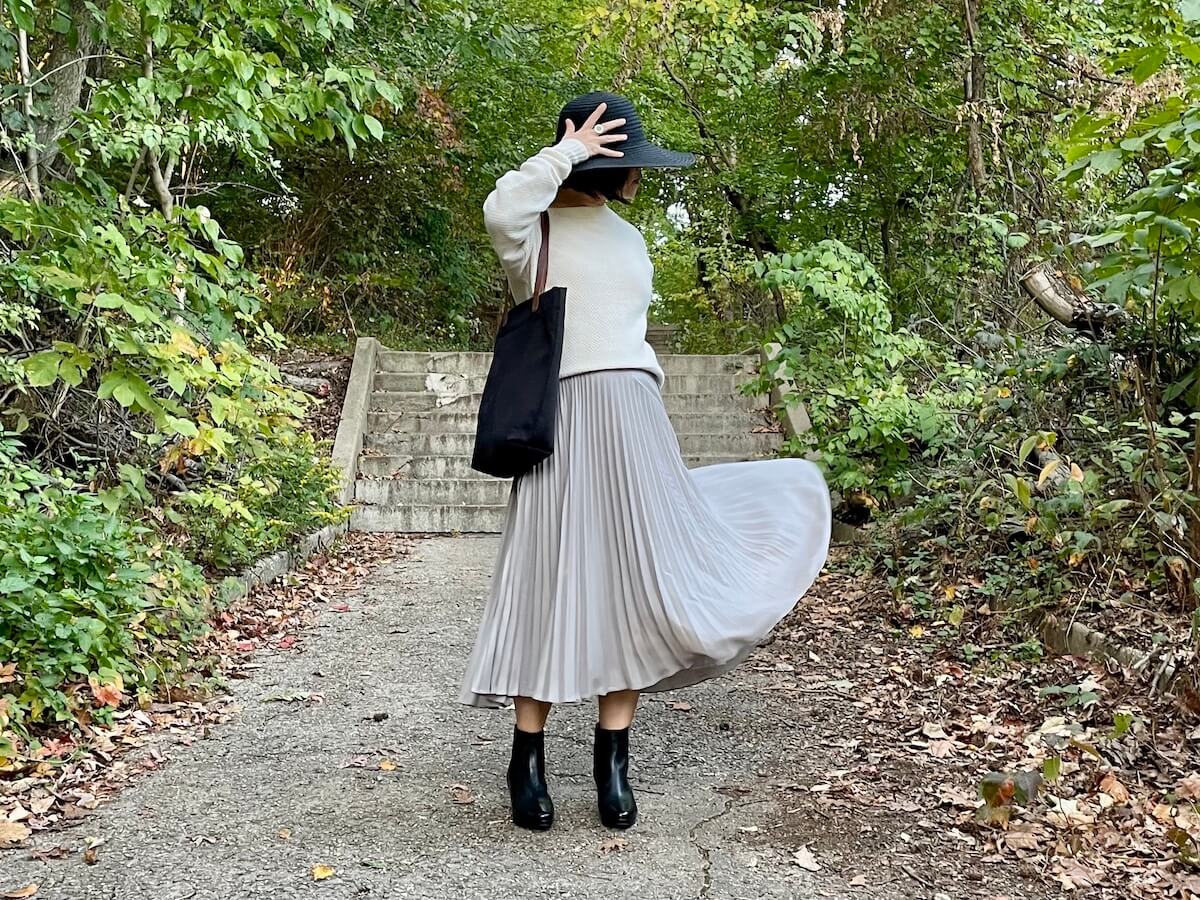 A person posting in front of steps in a park, wearing a white sweater, gray pleated midi skirt, and black booties and black hat. The skirt is in motion.