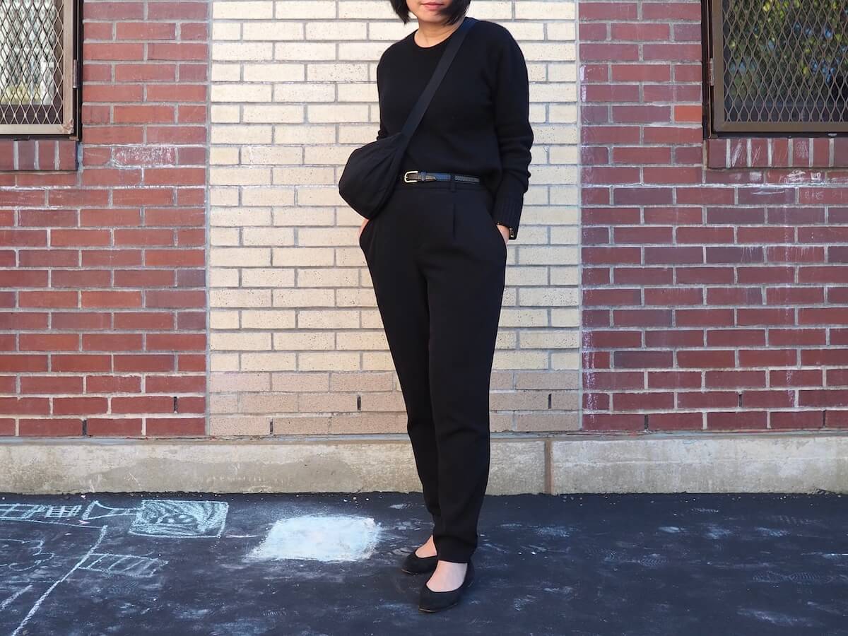 Me, a person with black hair, wearing an all-black outfit from sweater to pants, standing in front of a brick wall made of yellow and red bricks. My hands are in my pockets.