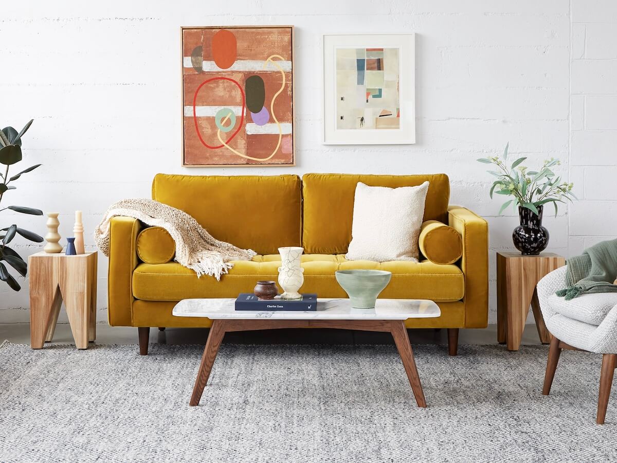 A yellow velvet sofa with tufted seat cushions. There is a marble top coffee in front of it and framed artwork behind it.