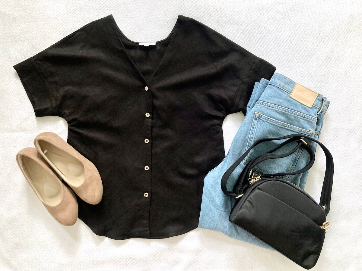 A flatlay of clothing items: black short sleeve button up, jeans, a black nylon bag, and brown wedge shoes.