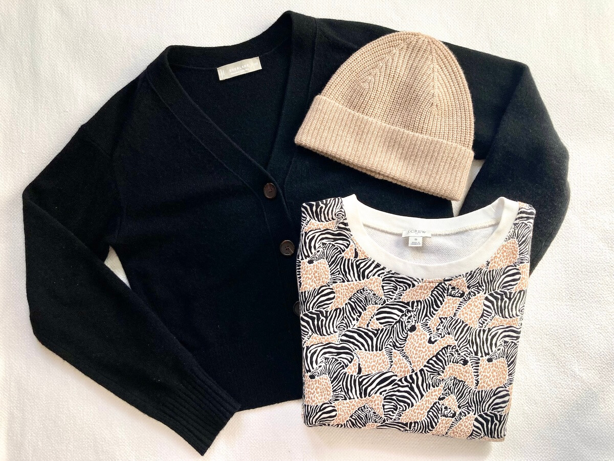 A flatlay of three items: a black cropped cardigan, a beige beanie, and a sweatshirt with a repeating zebra design.