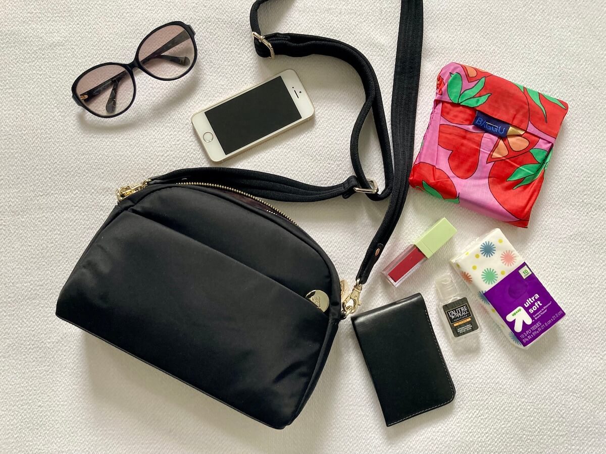Flatlay featuring a black crossbody bag, the Nouvelle, with items that fit in it, including wallet, sunglasses, reusable bag, and more.
