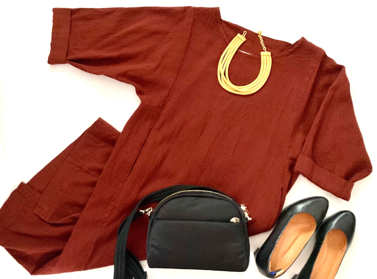 Flatlay of a maroon sack dress with black heels, a black dome-shaped purse, and a gold necklace.