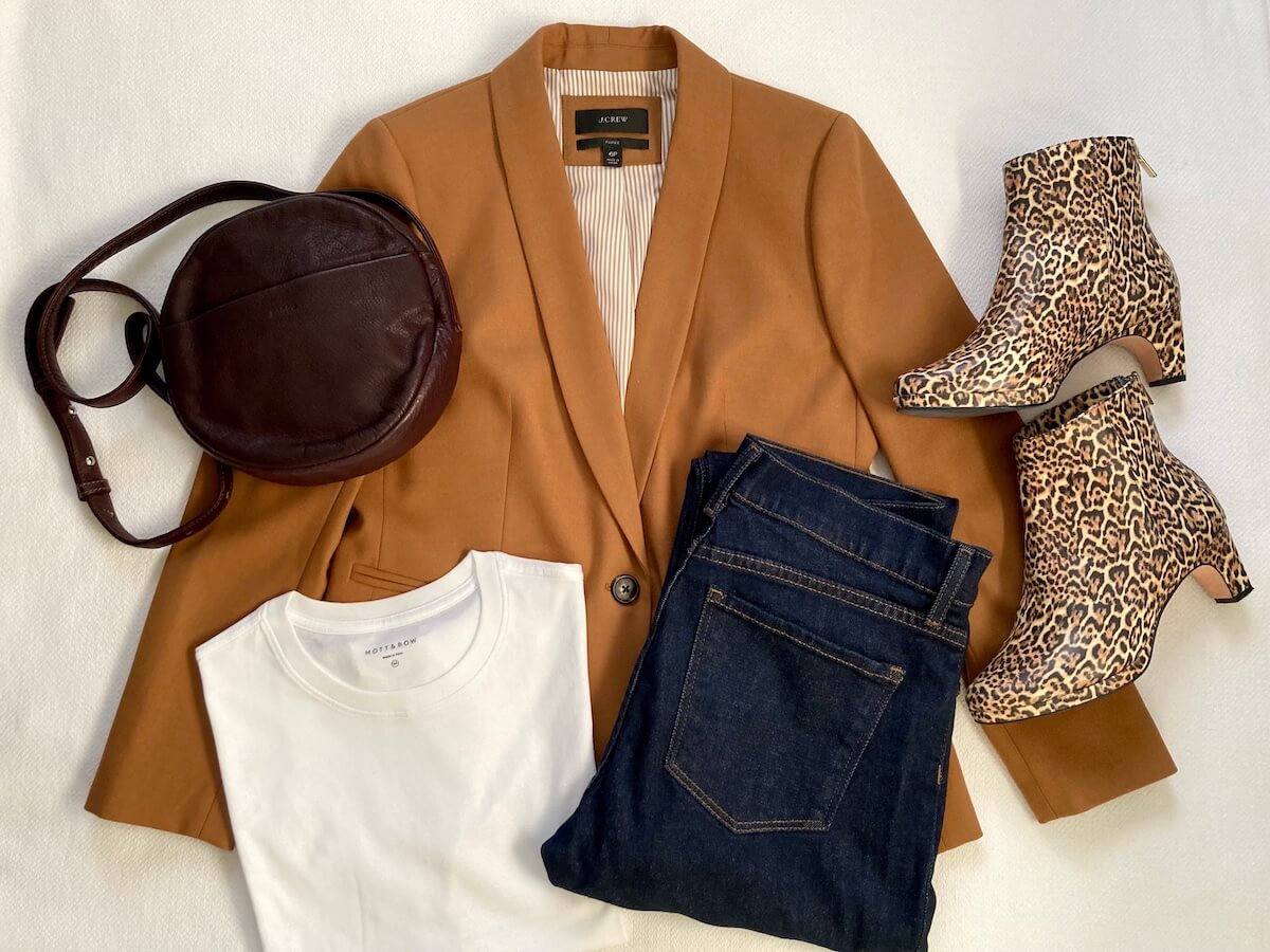A flatlay of an outfit consisting of a camel blazer, blue jeans, white t-shirt, leopard print booties, and a maroon circle purse.