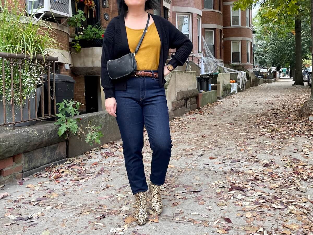A person wearing a mustard yellow shirt tucked into mom jeans with a black cardigan and leopard print bag. They have a hand on hip.