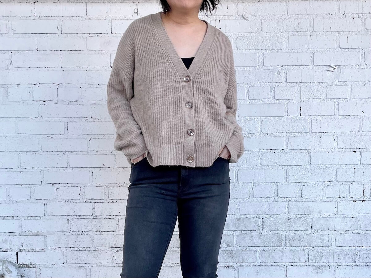 A person with dark hair stands in front of a brick wall wearing an oversized cardigan that is buttoned over skinny jeans.