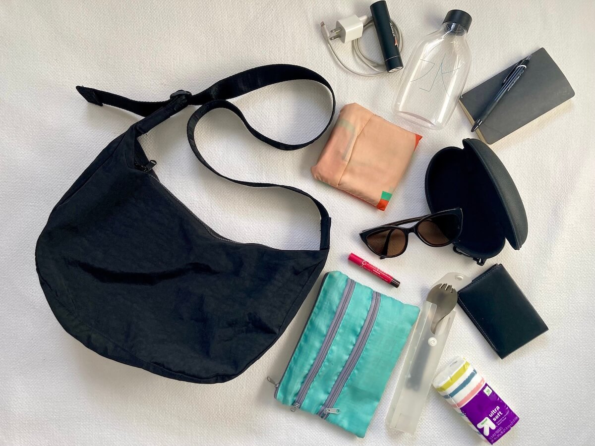 A flatlay showing a black crescent bag from Baggu and various items including a pouch, a notebook, sunglasses case, spork, tissues, charger, and more.
