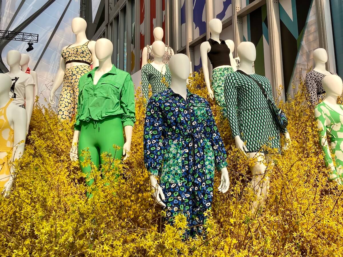 Mannequins wear clothing with bold prints in green, blue and yellow, among yellow plants.