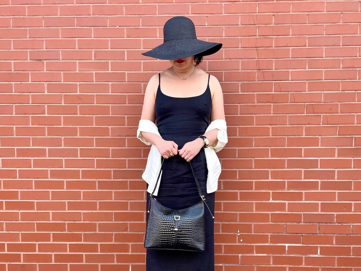 A person with dark hair stands in front of a red brick wall wearing a black dress with black hat and holding a black bag in both hands in front of them.