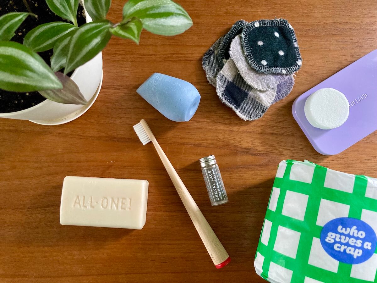 A flat lay of items arrange on a wooden surface including a bamboo toothbrush, glass container for floss, bar shampoo, Who Gives a Crap toilet paper, and more.