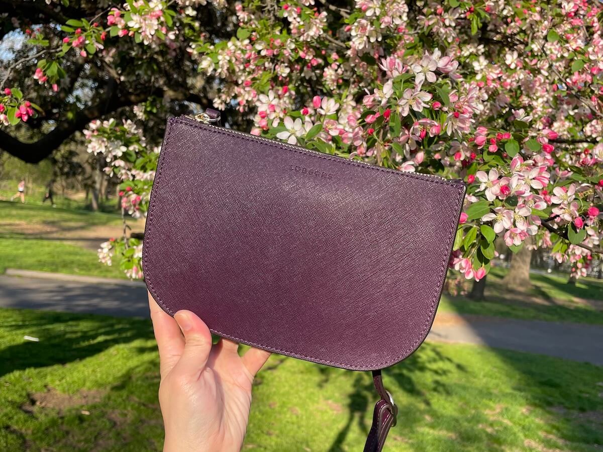 A hand holds up a Lo & Sons small bag with rounded bottom corners. Behind is a cherry blossom tree.