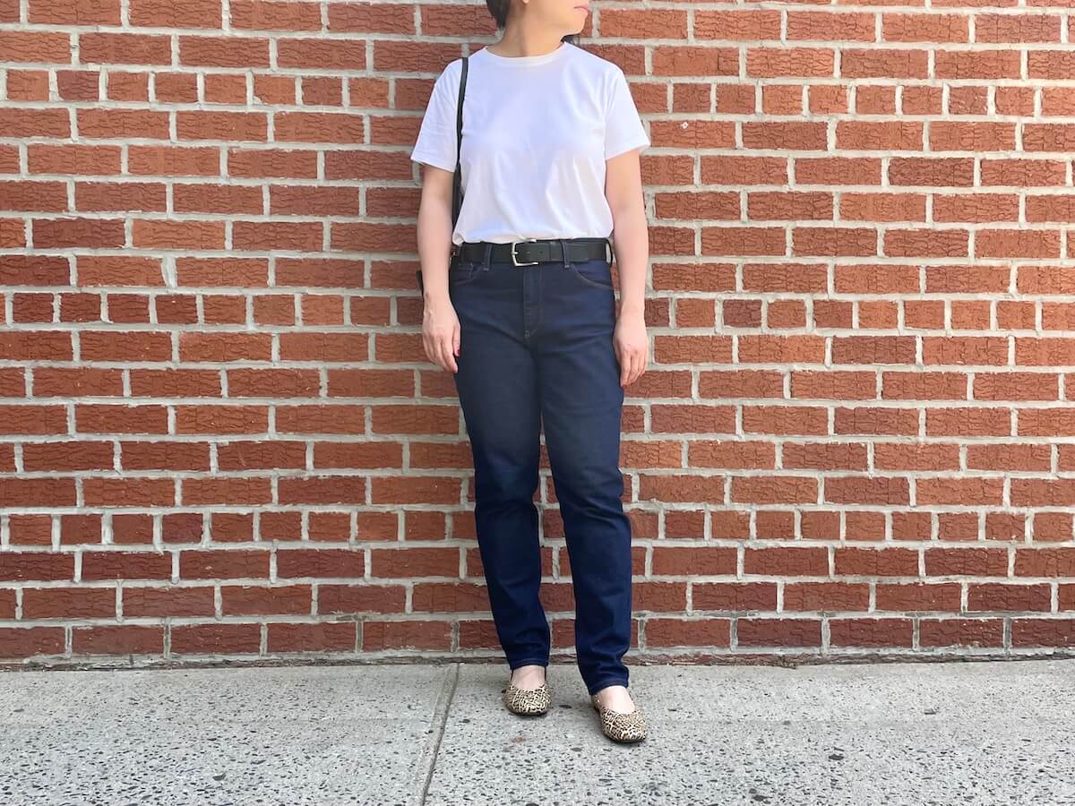 A person stands in front of a red brick wall wearing dark, slim jeans and white T-shirt.