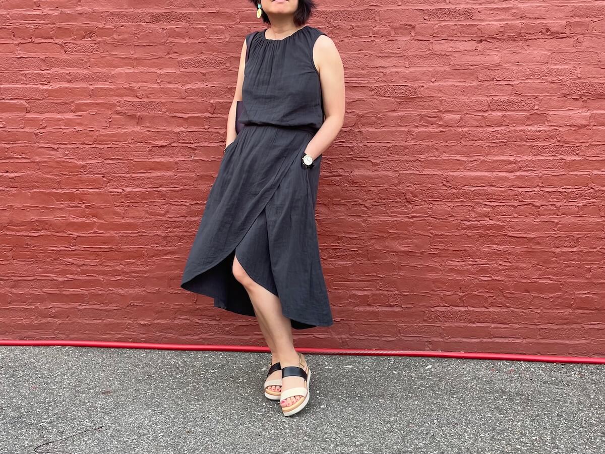 A person wearing a black sleeveless bubble top and a matching wrap skirt in front of a red wall. Their hands are in their pockets.