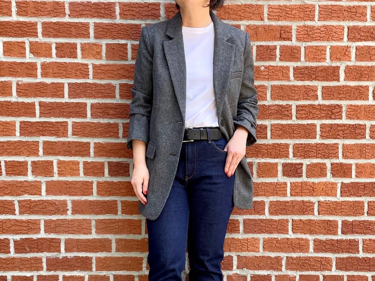 A person wearing a gray oversized blazer and jeans standing in front of a brick wall with one hand hooked in a jean pocket.