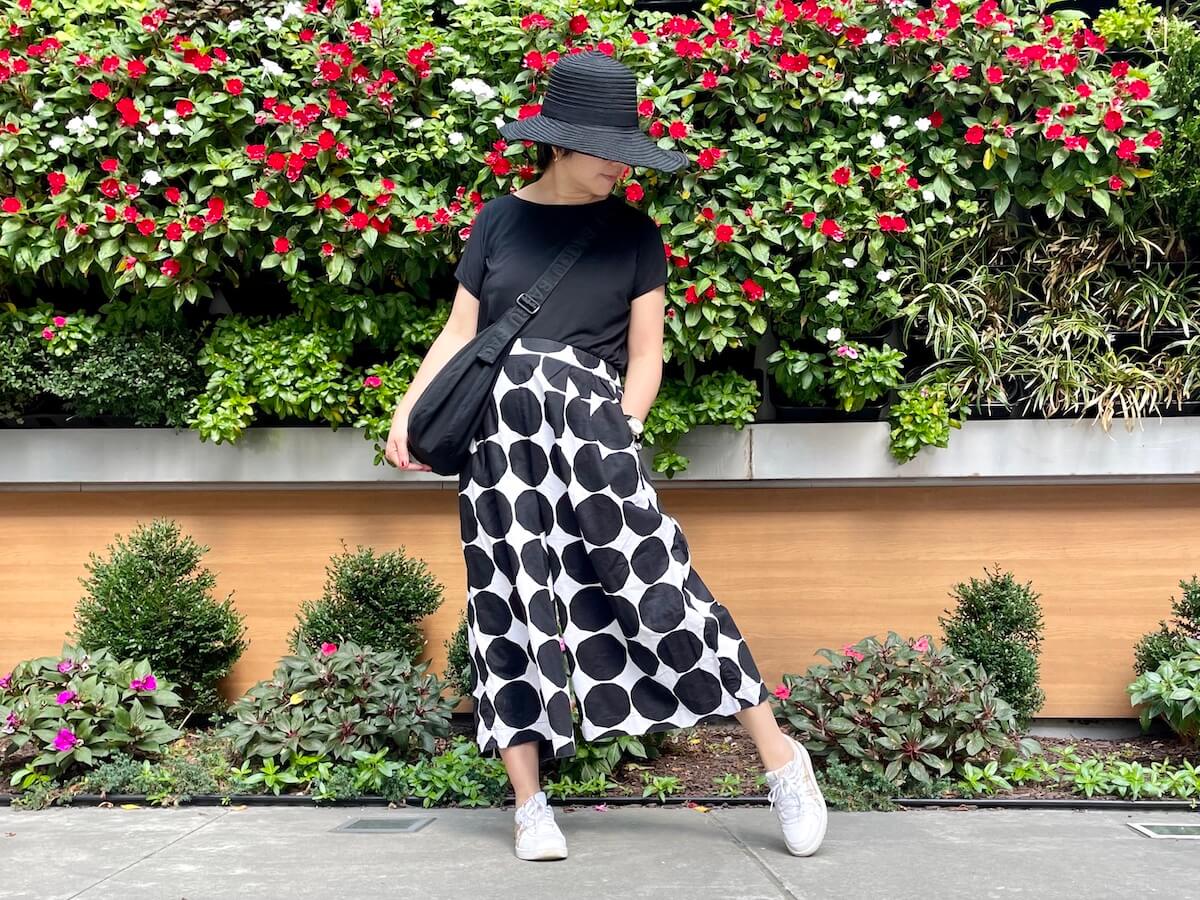 A person wearing a black tee and large dot print wide legged pants posing in front of a wall of vegetation