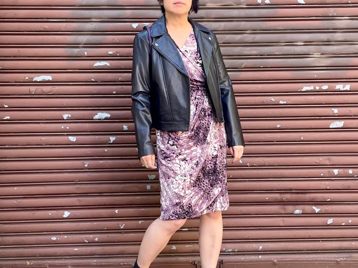 A person wearing a black leather moto jacket over a purple dress poses in front of a metal roll-down door.