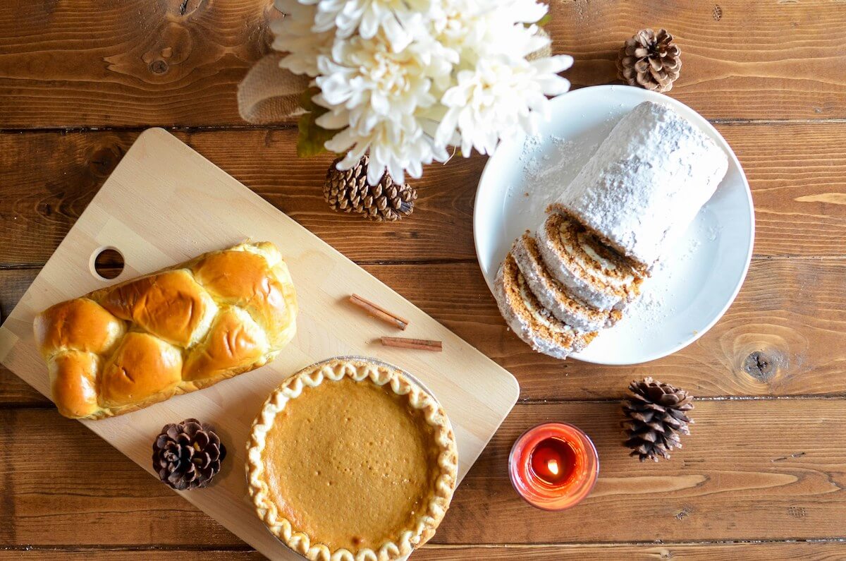 A table with desserts, viewed from above. There's pumpkin pie, bread, and a cake roll. There are pinecones, a candle, and flowers too.