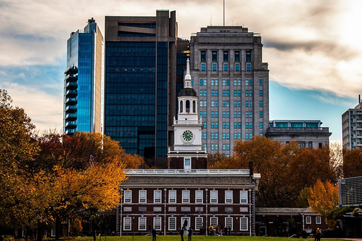 Independence Hall, a historic building with a spire, in front of modern buildings. There are orange trees surrounding it.