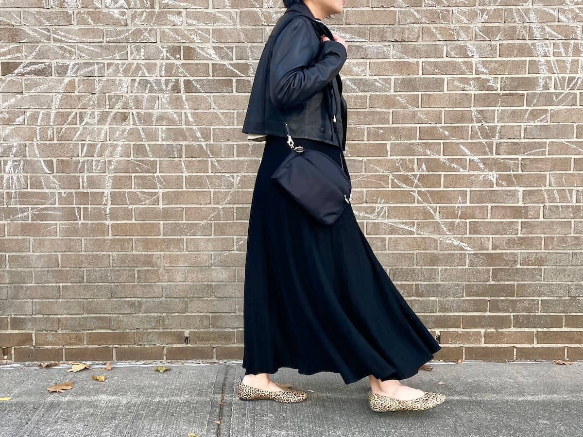 A person wearing a cropped black moto jacket and a black maxi skirt walks in front of a brick wall with chalking on it.