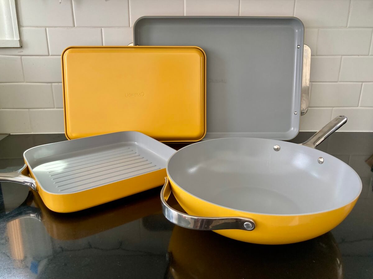 Two bake sheets, a square grill pan, and a stir fry pan, all in marigold yellow, from Caraway arranged on a counter.