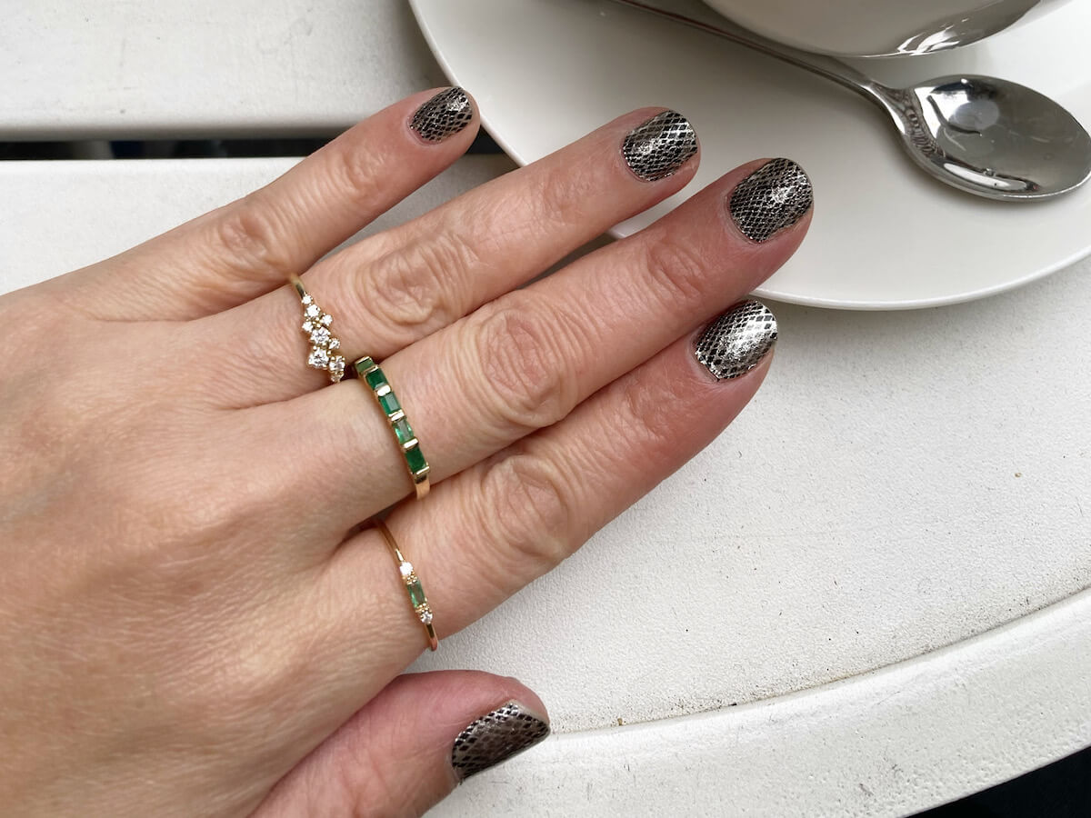 A hand with three rings, one of them diamond, two of them with emeralds, on a hand with snakeskin patterned nail polish.