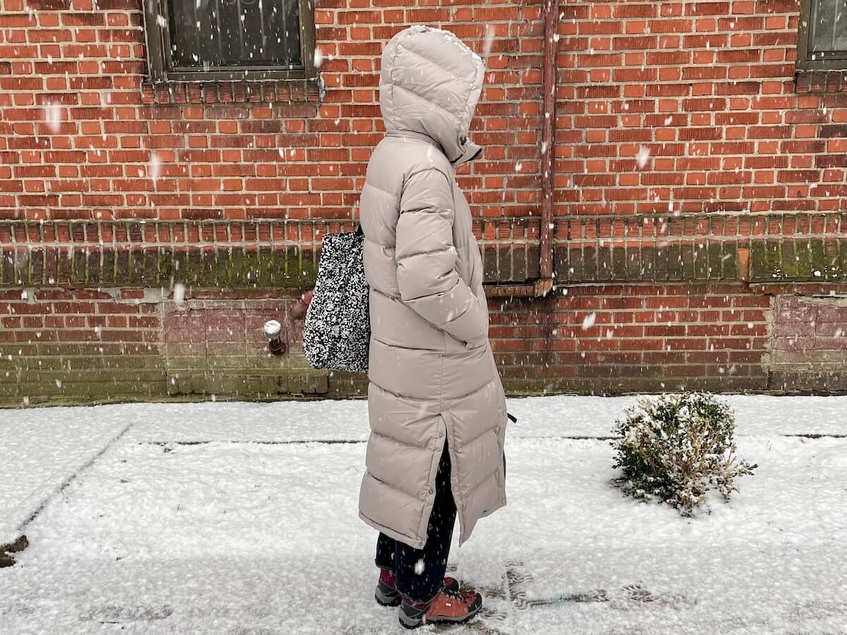 A person wearing a long puffer jacket with the hood up, standing in front of a red brick building. It is snowing.
