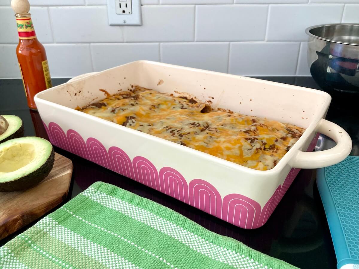 A Great Jones baking dish with a pink design on a counter, with enchiladas topped by melted cheese.