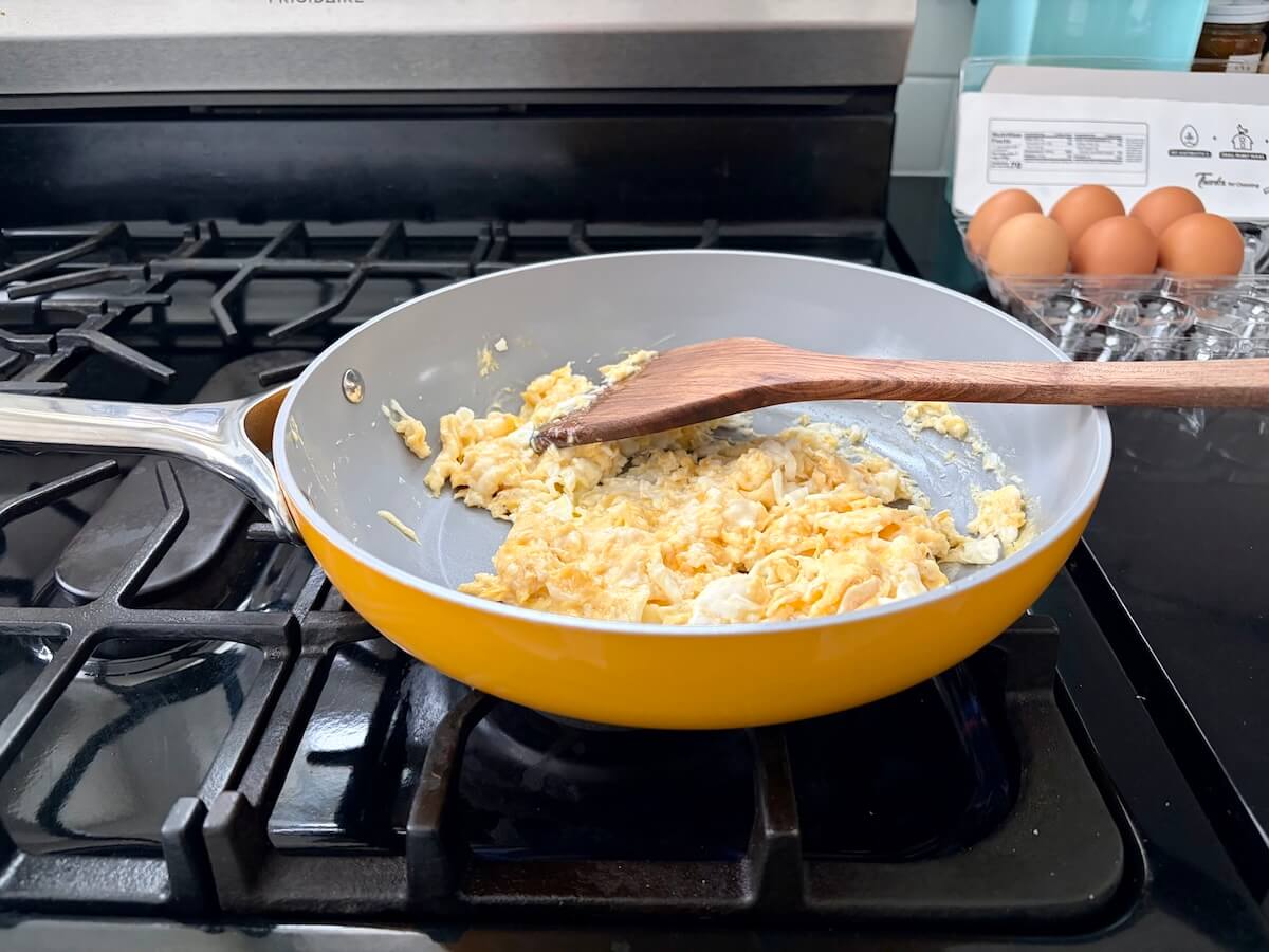 A yellow frypan on a stove with scrambled eggs. A box of eggs sits on the counter.