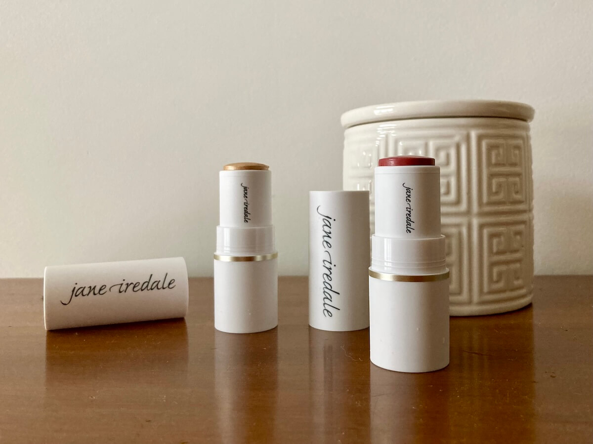 Two tubes of jane iredale products, which are open, on a wooden surface. There is a lidded white ceramic jar behind them.